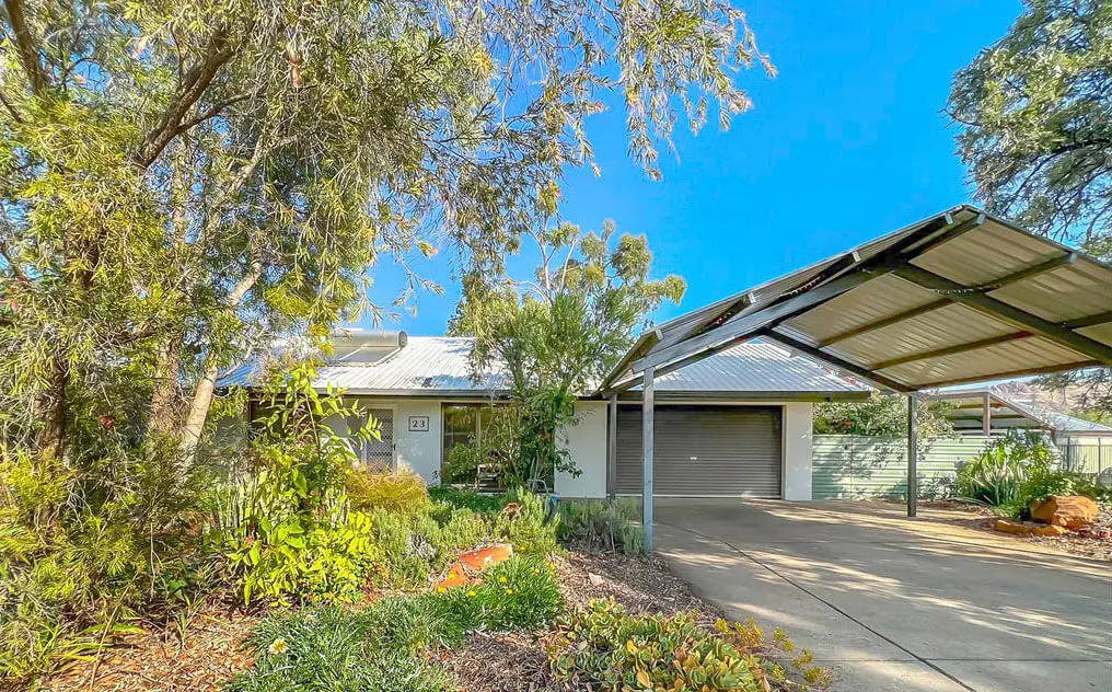Single-story house with a metal roof, surrounded by lush greenery. There is a covered carport on the right and a concrete driveway leading to a garage. Bright blue sky overhead.