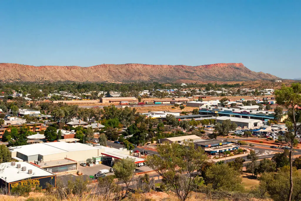A panoramic view of a small town with buildings and roads, including the Coe Industrial area, surrounded by trees and shrubland, set against a backdrop of red rocky hills under a clear blue sky.