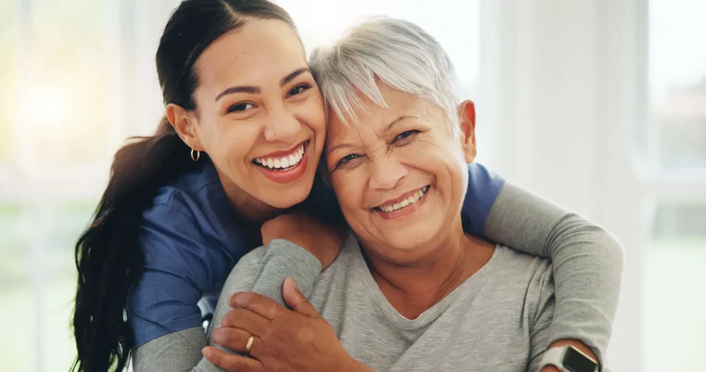 A younger woman hugs an older woman from behind as they both smile warmly at the camera, conveying happiness and affection in a bright indoor setting at Coe Industrial.