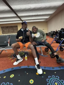 Two men sit and eat sandwiches in an airport waiting area. One wears an orange outfit and a cap, while the other wears a green hoodie and black pants. A walker is next to them on the right. The floor has a colorful patterned carpet.