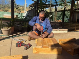 A man sits on a sunny patio using a power drill to work on a wooden project. Wooden planks, tools, and plants are around him, and a shaded structure and garden can be seen in the background.