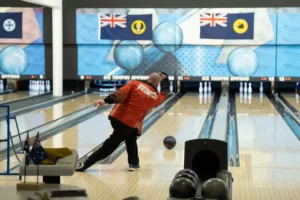 A person wearing a red shirt bowls at a ten-pin bowling alley. Two blue balls are visible on adjacent lanes, and pins are set up ahead. Flags hang above the lanes in the background. Bowling balls sit on a rack in the foreground.