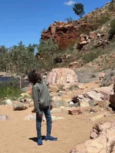 A person wearing a gray hoodie, jeans, and blue shoes stands on sandy ground surrounded by rocks and greenery, looking up toward the sky, with rocky hills and trees in the background under a clear blue sky.