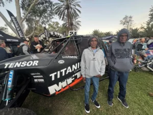 Two men stand in front of a large off-road racing vehicle with "Titanium" and "Tensor" logos at an outdoor event. Other people, palm trees, and banners are visible in the background.
